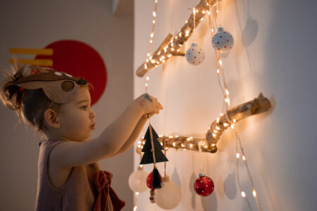 Ein kleines Kind mit einem Rentier-Maske hängt Weihnachtsbaumschmuck an einer beleuchteten Wanddekoration aus Ästen. Die Wand ist mit Lichtern und Weihnachtskugeln geschmückt, darunter eine schwarze Weihnachtsbaumfigur und eine rote Kugel.