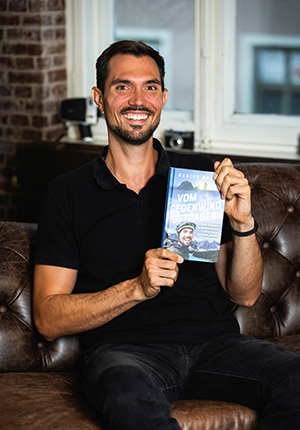 Ein Mann sitzt auf einem braunen Ledersofa und lächelt in die Kamera. Er trägt ein schwarzes T-Shirt und hält ein Buch mit dem Titel 'Vom Gegenwind getragen' in der Hand. Im Hintergrund sind Fenster und einige technische Geräte sichtbar.
