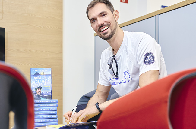 Ein Mann sitzt auf einem roten Sessel und lächelt, während er ein Buch signiert. Auf dem Tisch neben ihm stehen mehrere Exemplare des Buches "Vom Gegenwind getragen". Er trägt ein weißes T-Shirt mit einem Logo und Sonnenbrille auf dem Kopf. Der Hintergrund zeigt eine helle, moderne Büroumgebung.