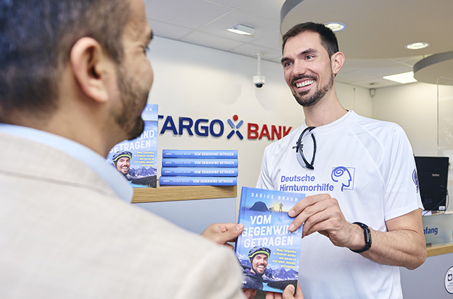 In der Bankfiliale von TARGOBANK steht ein lächelnder Mann in einem weißen Shirt, der ein Buch mit dem Titel "VOM GEGENWIND GETRAGEN" hält. Er spricht mit einem anderen Mann, der einen Anzug trägt. Im Hintergrund sind weitere Exemplare des Buches auf einem Regal zu sehen, sowie das Logo der Deutschen Hirntumorhilfe.