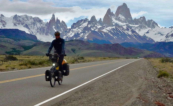 Ein Radfahrer fährt auf einer Straße durch eine beeindruckende Berglandschaft mit schneebedeckten Gipfeln im Hintergrund. Die Szenerie zeigt eine weite, grüne Landschaft mit Bergen, die majestätisch in den Himmel ragen. Der Radfahrer trägt einen Helm und hat Gepäck an seinem Fahrrad befestigt.