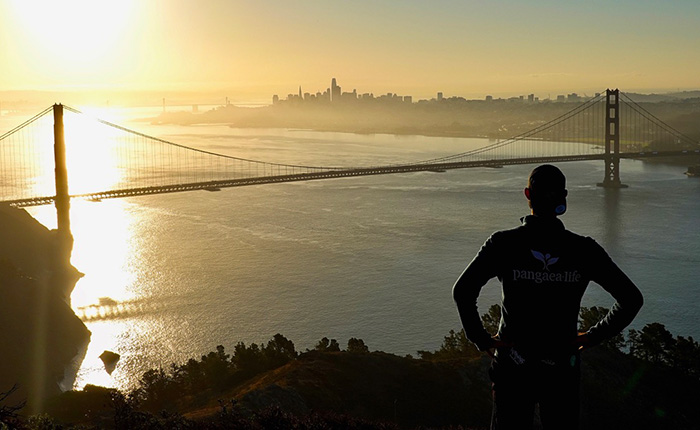 Eine Person steht auf einem Hügel mit Blick auf die Golden Gate Bridge und die Skyline von San Francisco im Hintergrund. Die Sonne geht auf und reflektiert auf dem Wasser, während die Stadt in der Dämmerung sichtbar ist. Die Person trägt ein schwarzes Shirt mit dem Logo von Pangaea Life.