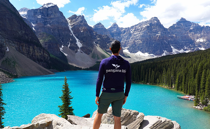 Ein Mann steht auf einem Felsen am Ufer eines turquoisefarbenen Sees, umgeben von hohen Bergen mit schneebedeckten Spitzen. Er trägt einen dunkelblauen Kapuzenpullover mit dem Logo 'pangea.life' und shorts. Im Hintergrund sind dichte Wälder und einige Boote am Ufer sichtbar. Der Himmel ist klar mit wenigen Wolken.