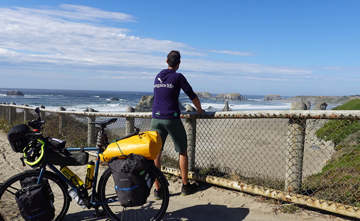 Ein Mann steht an einem Geländer und schaut auf die Küste. Er trägt ein lila Sweatshirt mit dem Aufdruck 'BaysceneLife' und grüne Shorts. Neben ihm steht ein Fahrrad mit Taschen, und im Hintergrund sind Wellen und Felsen am Strand sichtbar.