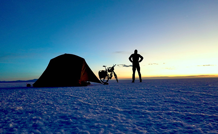 Ein Mann steht in der Silhouette vor einem Zelt und einem Fahrrad auf einer weiten, weißen Salzebene. Der Himmel zeigt Farbnuancen von Blau und Orange während des Sonnenuntergangs.