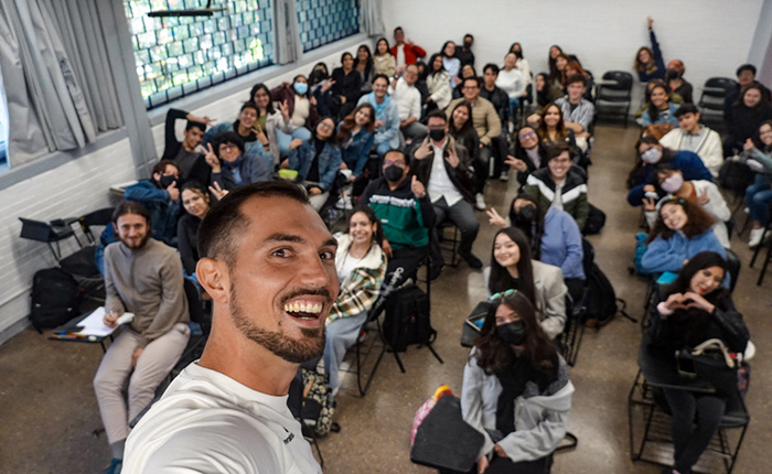 Ein Selbstporträt eines Mannes in einem Klassenraum mit vielen Studierenden im Hintergrund. Der Mann lächelt in die Kamera, während die Gruppe von etwa 40 bis 50 Personen, die größtenteils lässige Kleidung tragen, in unterschiedlichen Posen für das Foto posiert, einige mit Handzeichen oder lächelnd. Der Raum hat helle Wände und große Fenster, durch die Tageslicht strömt.