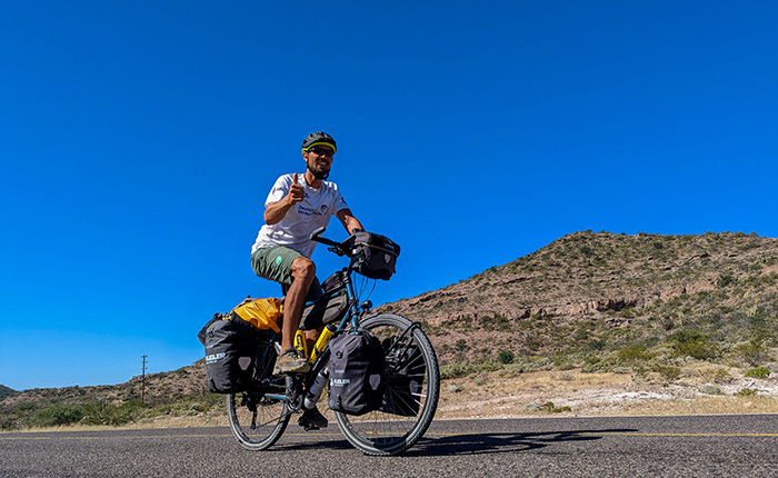 Ein Radfahrer auf einem Fahrrad fährt auf einer Straße. Er trägt ein weißes T-Shirt und grüne Shorts, mit einem Helm auf dem Kopf. Der Radfahrer zeigt mit dem Daumen nach oben. Im Hintergrund ist eine hügelige Landschaft mit einem klaren blauen Himmel sichtbar. Das Fahrrad hat zwei große Gepäcktaschen an den Seiten.