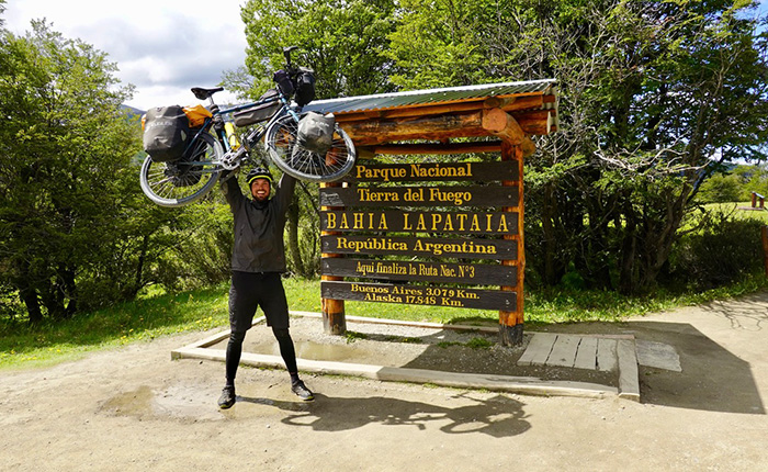 Ein Mann hebt sein Fahrrad über den Kopf, während er vor einem Holzschild steht, das 'Bahía Lapataia' und 'Parque Nacional Tierra del Fuego' anzeigt. Das Schild enthält Informationen über die Entfernung nach Buenos Aires und Alaska. Im Hintergrund sind Bäume und eine grüne Landschaft sichtbar.