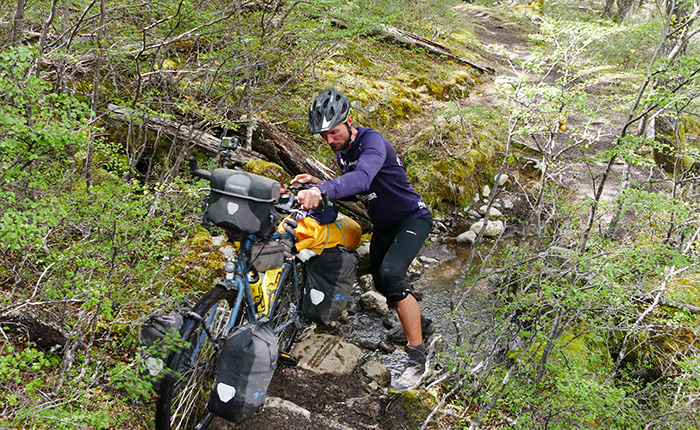 Ein Radfahrer mit einem Helm kämpft, um sein Fahrrad über einen steinigen, bewachsenen Pfad zu schieben. Das Fahrrad ist mit Gepäcktaschen beladen. Um ihn herum sind grüne Pflanzen und Bäume. Der Weg ist uneben und führt durch ein kleines Wasserbett.