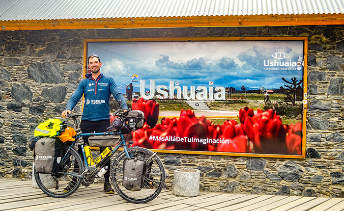 Ein Mann steht neben einem Fahrrad mit Gepäck vor einem Schild mit der Aufschrift "Ushuaia". Im Hintergrund sind rote Blumen und eine Berglandschaft zu sehen. Der Mann trägt ein blaues Sweatshirt mit Logos und lächelt.