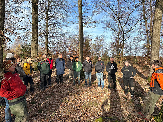 Eine Gruppe von etwa 15 Personen steht in einem Waldgebiet mit Laub auf dem Boden. Die Menschen tragen verschiedene Kleidungsstücke, darunter Jacken und Sweatshirts. Einige haben Rucksäcke, und die Sonne scheint durch die Bäume. Im Hintergrund sind weitere Bäume und Sträucher sichtbar.