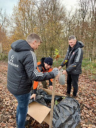 Drei Personen in Sportjacken arbeiten gemeinsam im Freien, um Pflanzen aus einer schwarzen Plastiktüte zu entnehmen. Der Boden ist mit Laub bedeckt.