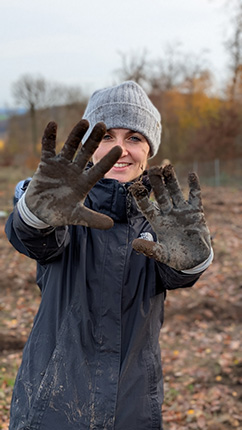 Eine Person trägt eine graue Mütze und eine dunkle Jacke. Ihre Hände sind schmutzig und sie hält sie nach vorne, während sie sich in einer ländlichen Umgebung mit herbstlichen Bäumen im Hintergrund befindet.