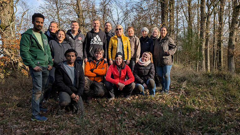 Eine Gruppe von 14 Personen steht in einem Waldgebiet. Sie tragen verschiedene Freizeitkleidung, darunter Jacken, Hoodies und Sportkleidung. Die Umgebung zeigt Bäume und Gras. Es ist Tageslicht zu sehen.