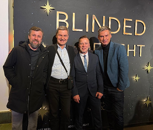 Vier Männer posieren vor einer Wand mit dem Schriftzug 'BLINDED BY LIGHT'. Sie tragen formelle Kleidung und haben unterschiedliche Frisuren.