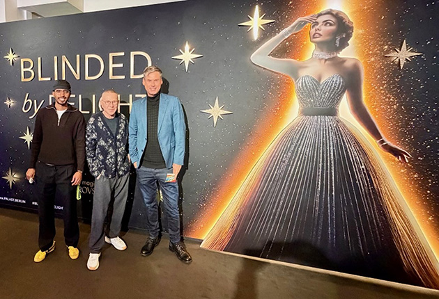 Drei Personen stehen vor einem großen Banner mit dem Text "BLINDED by DELIGHT" und einer glamourösen Figur in einem glitzernden Kleid. Die Personen tragen verschiedene Outfits und posieren für das Bild.