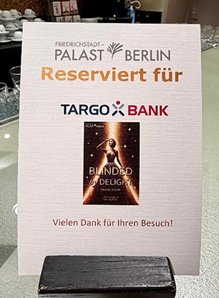 Ein Schild steht auf einem Tisch mit dem Text: 'Reserviert für TARGO BANK'. Darunter befindet sich ein Bild des Posters für die Show 'BLINDED by DELIGHT' mit dem Hinweis: 'Vielen Dank für Ihren Besuch!'.