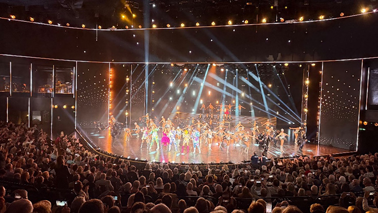 Eine lebhafte Bühnenaufführung mit vielen Tänzern in bunten Kostümen auf einer hell beleuchteten Bühne, umgeben von einem begeisterten Publikum.