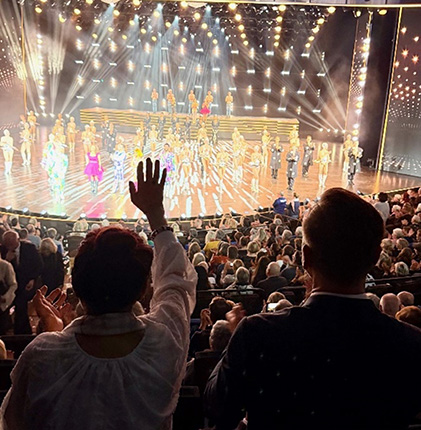 Zuschauer applaudieren begeistert bei einer Bühnenaufführung mit vielen Tänzern in bunten Kostümen und hellen Lichtern.