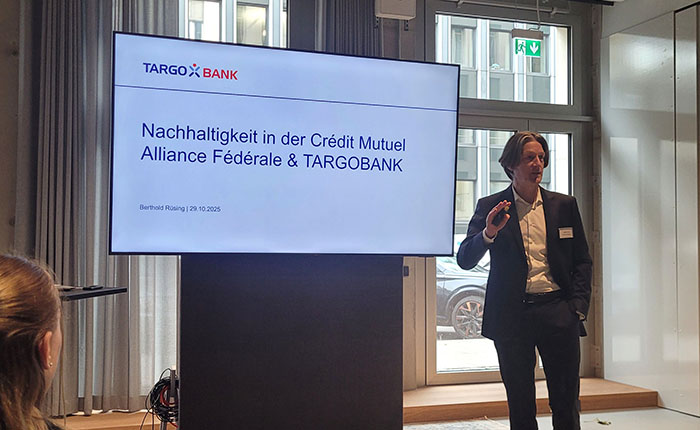 Ein Mann steht vor einem Bildschirm mit dem Logo von TARGOBANK und einer Präsentation über Nachhaltigkeit. Die Folie zeigt den Titel "Nachhaltigkeit in der Crédit Mutuel Alliance Fédérale & TARGOBANK" sowie das Datum "29.10.2025". Der Mann trägt einen schwarzen Anzug und hält einen Arm hoch, während er spricht. Im Hintergrund sind Fenster und eine andere Person sichtbar.