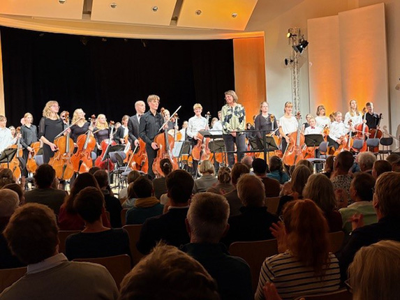 Ein Orchester mit mehreren Musikern, die auf der Bühne stehen. Im Vordergrund applaudiert das Publikum. Die Musiker spielen Celli und tragen schwarze oder weiße Kleidung. Im Hintergrund sind weitere Orchestermitglieder sichtbar, die sich auf ihren Instrumenten vorbereiten.