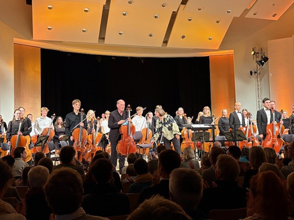 Eine Orchestervorstellung mit Musikern, die vor dem Publikum auf der Bühne stehen. Im Vordergrund sind mehrere Cellisten zu sehen, die applaudieren. Die Bühne ist gut beleuchtet mit orangefarbenen Akzenten. Das Publikum sitzt in einem abgedunkelten Raum und applaudiert.