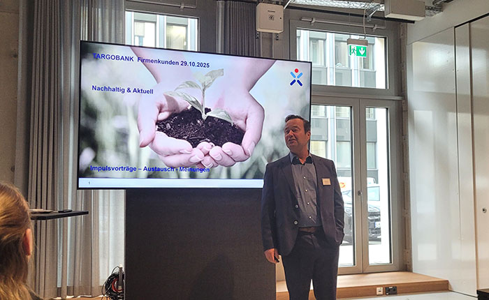 Ein Mann steht an einem Präsentationsbildschirm, der ein Bild von Händen mit Erde und einer kleinen Pflanze zeigt. Der Bildschirm zeigt den Text "TARGOBANK Firmenkunden 29.10.2025 Nachhaltig & Aktuell Impulsvorträge - Austausch - Meinungen". Der Raum hat große Fenster und einen modernen Look.