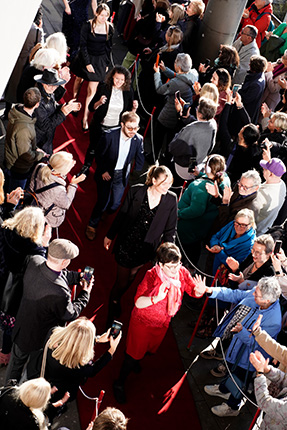 Eine Menschenmenge applaudiert auf einem roten Teppich, während mehrere Personen, darunter eine Frau in rotem Kleid, vorbeigehen.