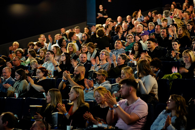 Eine große Menschenmenge sitzt in einem Raum und applaudiert. Die Zuschauer sind in verschiedenen Positionen und Gesichtern sichtbar, alle applaudieren enthusiastisch. Einige Personen tragen lässige Kleidung und es sind Getränke auf den Sitzen sichtbar. Die Atmosphäre ist lebhaft und energisch.