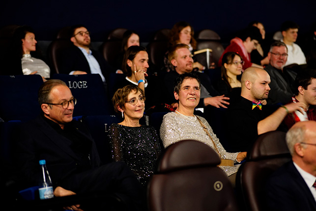 Eine Menschenmenge sitzt in einem dunklen Raum mit Stühlen. Einige Personen tragen formelle Kleidung, während andere lässiger gekleidet sind. Im Vordergrund sind zwei Personen sichtbar, die sich unterhalten. Auf dem Tisch vor ihnen steht eine Wasserflasche.