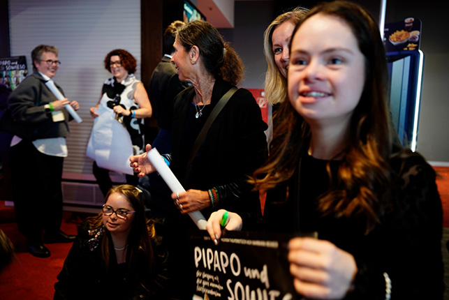 Eine Gruppe von Menschen steht in einem Innenraum. Einige Personen halten Papierrollen und ein Schild mit der Aufschrift "PIPAPO und SOHNES". Die Szene ist lebhaft und zeigt Interaktion unter den Anwesenden.