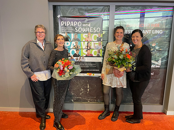 Vier Personen stehen vor einem Plakat mit der Aufschrift "PIPAPO und SOWIESO". Zwei halten Blumensträuße. Die Umgebung ist hell erleuchtet.