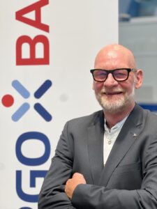 Eine Person in einem grauen Anzug steht mit verschränkten Armen vor einem Banner mit dem Wort 'BA'.