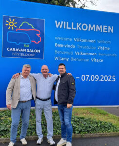 Drei Männer stehen vor einem großen blauen Banner mit dem Text 'WILLKOMMEN' und Informationen zum Caravan Salon Düsseldorf, der am 07.09.2025 stattfindet.