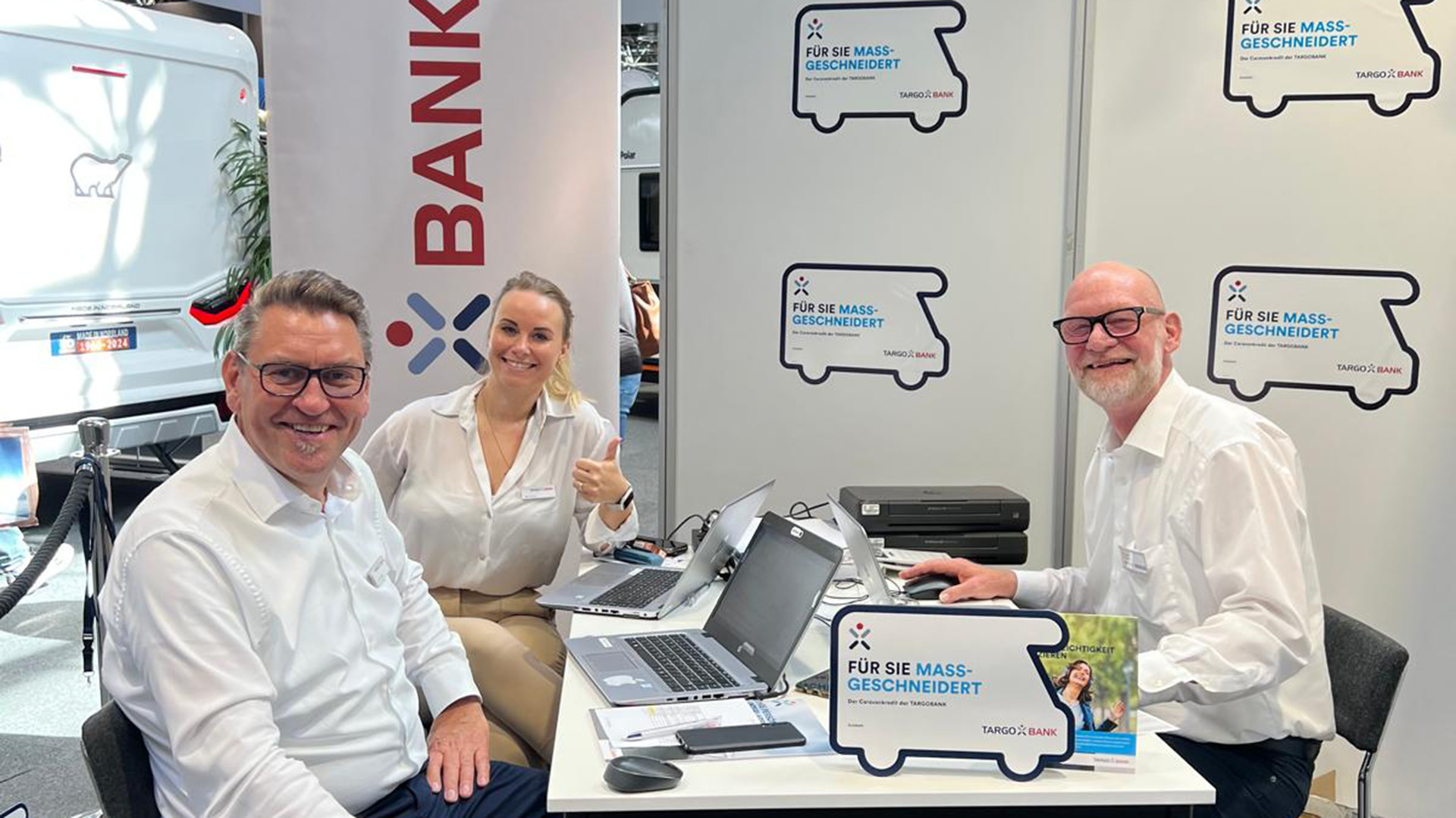 Drei Personen am Tisch mit Laptops, ein Banner mit dem Schriftzug 'BANK' und zwei Plakate der TARGOBANK mit dem Text 'FÜR SIE MASS-GESCHNEIDERT'.