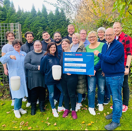 Eine Gruppe von Menschen steht im Freien auf einer Wiese, umgeben von Bäumen. Sie halten ein großes, blaues Plakat mit der Aufschrift "2000 €" und "TARGOBANK". Einige Personen halten einen weißen Behälter in der Hand.