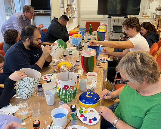 Eine Gruppe von Personen sitzt an einem Tisch und malt farbige Töpfe. Auf dem Tisch sind Pinsel, Farbtöpfe und Wassergläser verteilt.