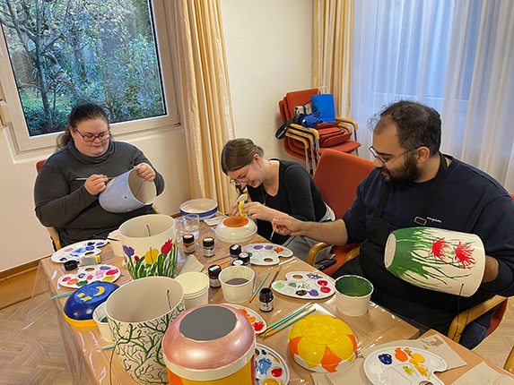 Drei Personen sitzen an einem Tisch und malen Töpfe. Auf dem Tisch sind verschiedene Pinsel, Farben und bemalte Töpfe. Im Hintergrund sieht man einen Fenster mit Vorhängen und einige Stühle.