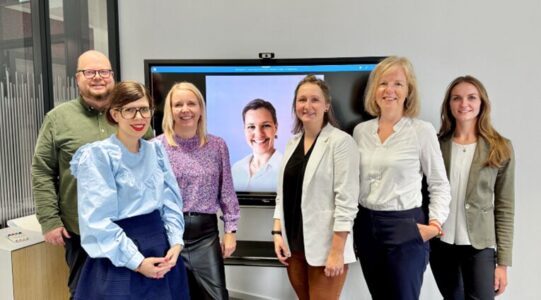 Eine Gruppe von sechs Personen steht in einem modernen Büro vor einem Bildschirm. Die Personen tragen formelle Kleidung und lächeln. Der Bildschirm zeigt ein Bild von einer weiteren Person.