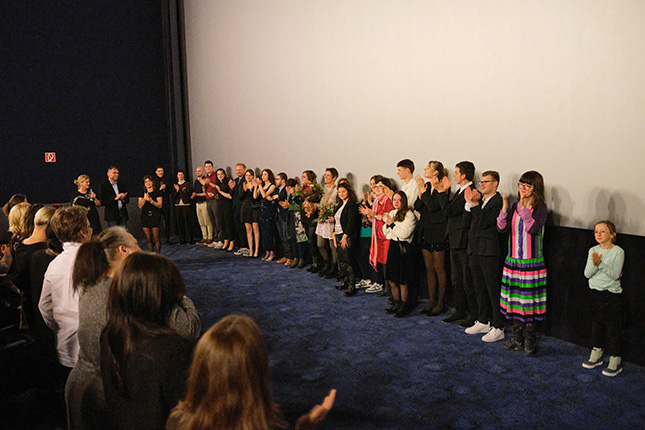 Eine Gruppe von Menschen steht auf einer Bühne in einem Kino und applaudiert. Die Anwesenden sind unterschiedlich gekleidet, einige tragen formelle Kleidung, während andere lässige Outfits haben. Im Vordergrund sind Zuschauer zu sehen, die ebenfalls applaudieren. Der Hintergrund zeigt eine große weiße Leinwand.