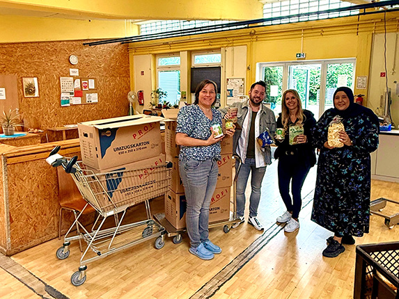 Vier Personen stehen in einem Raum mit großen Umzugskartons und einem Einkaufswagen. Sie halten Lebensmittel in den Händen.