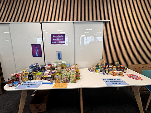 Ein Tisch mit verschiedenen Lebensmitteln, darunter Gläser, Dosen und Verpackungen, präsentiert in einem Raum. Zwei Plakate informieren über die Sammlung von Lebensmitteln vom 18.08. bis 22.08.2023.