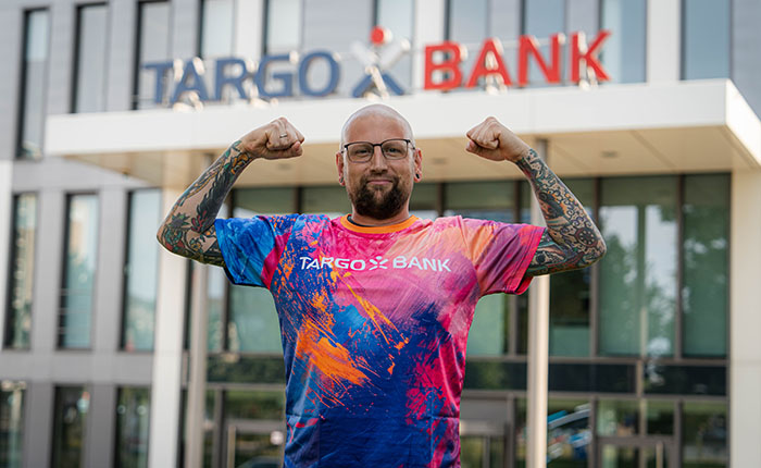 Ein Mann mit Glatze und Brille steht vor einem modernen Gebäude mit dem Schriftzug 'TARGO BANK'. Er trägt ein buntes T-Shirt mit dem Logo der Bank und hebt seine Arme stolz in die Luft.