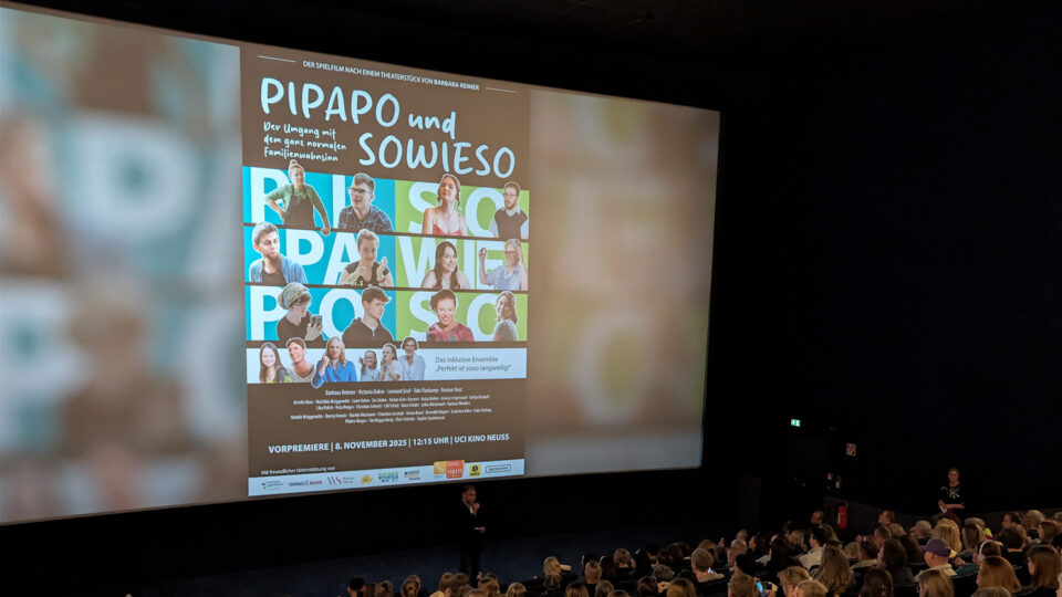 Das Bild zeigt eine Kinovorführung mit einer großen Menschenmenge. Auf der Leinwand ist das Plakat für den Film "PIPAPO und SOWIESO" zu sehen, das Details zur Premiere am 8. November 2025 enthält. Das Plakat zeigt mehrere Personen in verschiedenen Szenen, die den Titel des Films umgeben. Der Raum ist dunkel, und die Zuschauer scheinen auf die Präsentation zu achten.