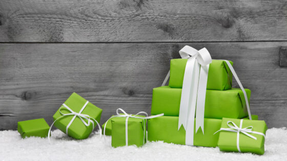 Eine Ansammlung von grünen Geschenken mit weißen Schleifen auf einem schneebedeckten Boden vor einer grauen Holzwand.