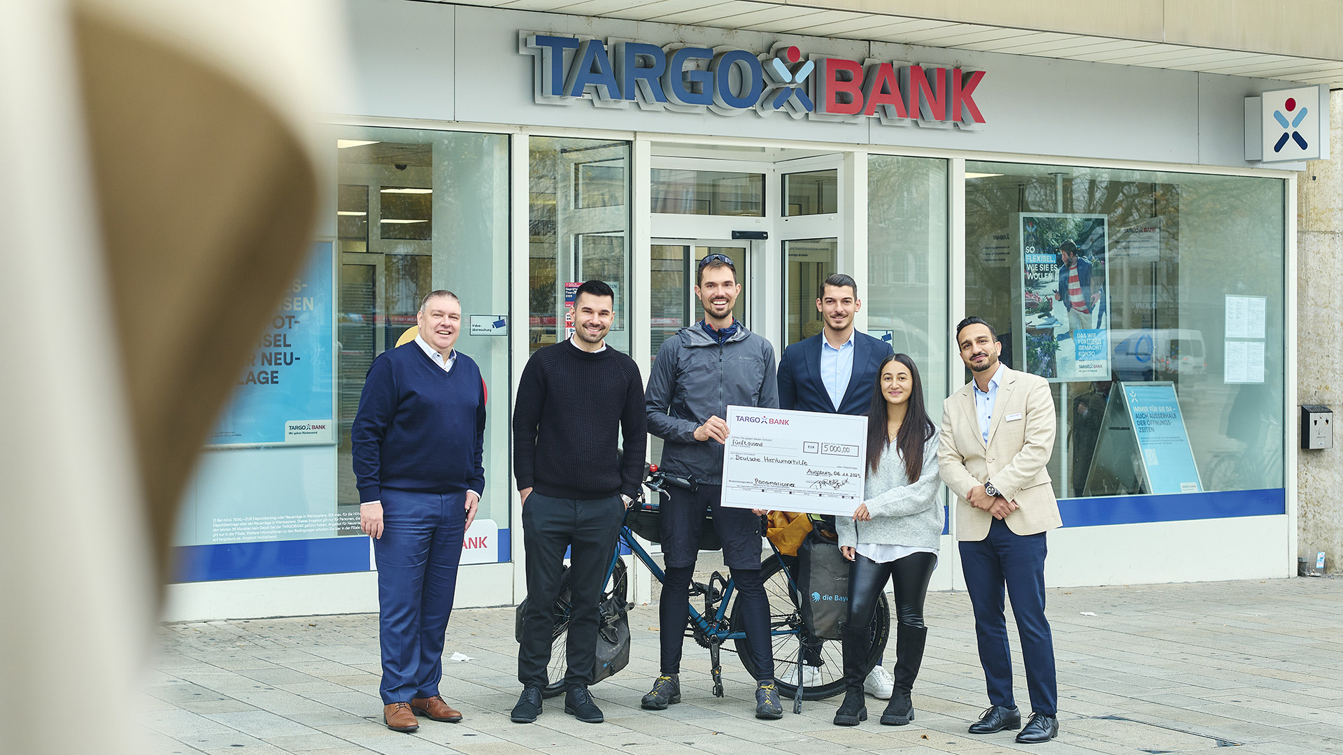 Eine Gruppe von fünf Personen steht vor einer TARGOBANK-Filiale. Sie halten einen großen Scheck über 600,00 Euro. Ein Mann in Sportkleidung mit einem Fahrrad ist in der Mitte. Die anderen Personen tragen formelle Kleidung. Im Hintergrund ist die Filiale mit einem blauen und weißen Logo sichtbar.