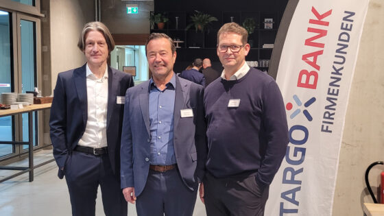 Drei Männer stehen in einem modernen Raum mit Regalen im Hintergrund. Links trägt der erste Mann einen schwarzen Anzug und ein weißes Hemd. In der Mitte steht ein Mann in einem blauen Anzug mit einem offenen Kragen und lächelt. Der dritte Mann rechts trägt einen dunklen Pullover und eine Brille. Im Hintergrund ist ein Banner der Stargo Bank mit dem Schriftzug 'Firmenkunden' zu sehen.