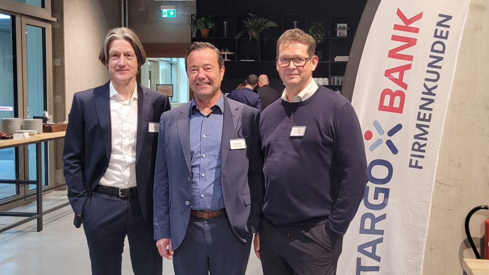 Drei Männer stehen in einem modernen Raum mit Regalen im Hintergrund. Links trägt der erste Mann einen schwarzen Anzug und ein weißes Hemd. In der Mitte steht ein Mann in einem blauen Anzug mit einem offenen Kragen und lächelt. Der dritte Mann rechts trägt einen dunklen Pullover und eine Brille. Im Hintergrund ist ein Banner der Stargo Bank mit dem Schriftzug 'Firmenkunden' zu sehen.