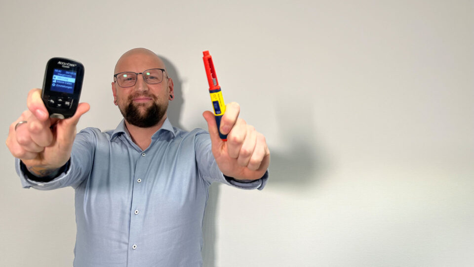 Ein Mann mit Glatze und Brille hält in jeder Hand ein medizinisches Gerät. In der linken Hand zeigt er ein Accu-Chek Blutzuckermessgerät und in der rechten Hand einen Injektor in Rot und Gelb. Der Hintergrund ist hell und neutral.