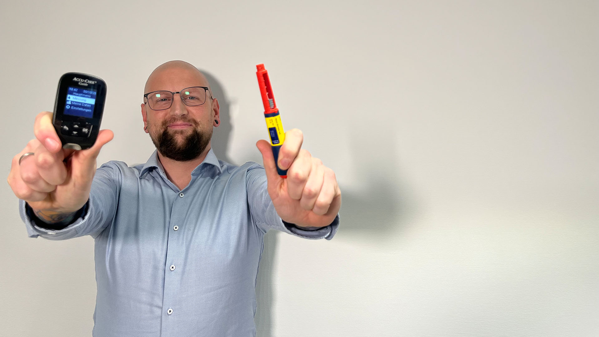 Ein Mann mit Glatze und Brille hält in jeder Hand ein medizinisches Gerät. In der linken Hand zeigt er ein Accu-Chek Blutzuckermessgerät und in der rechten Hand einen Injektor in Rot und Gelb. Der Hintergrund ist hell und neutral.